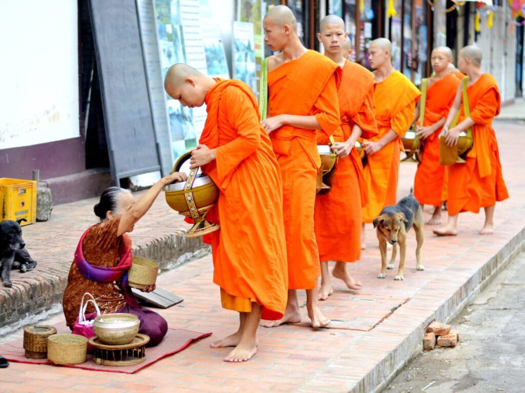 Monks in orange robes walking on the sidewalk receiving alms from an elderly woman